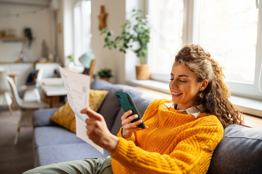 Woman sat on sofa looking at phone whilst holding a bill