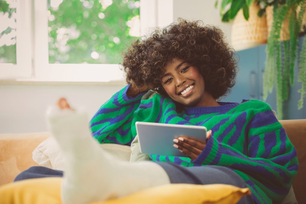 Afro American young woman wearing sweater sitting on sofa at home with broken leg in a plaster cast, holding digital tablet in hand and smiling at camera because she has an income protection policy