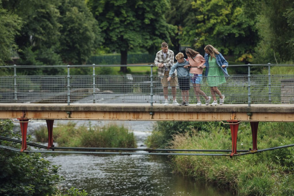 Wide shot of a family of four standing together on a bridge. The family consists of a mother and father with their son and daughter. The bridge is across a river located in Morpeth in the North East of England. They are looking down at the water.