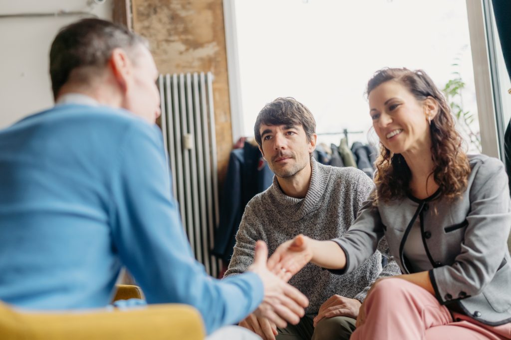 Three professionals engage warmly in a casual office environment, highlighting a friendly handshake. Perfect depiction of modern collaborative work culture.