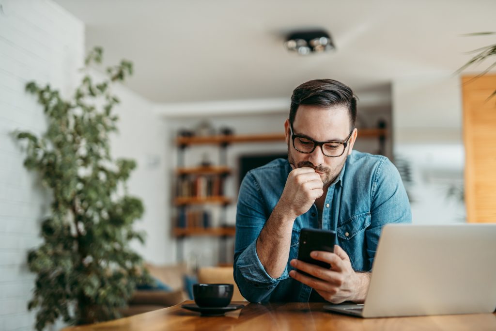Portrait of a puzzled man looking at smart phone, sitting at table at home.