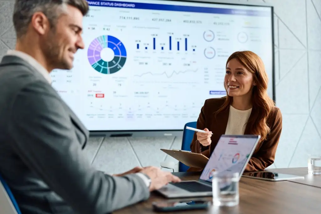 Two business professionals in a modern office looking at data visualizations on a large screen and laptop, discussing financial reports and strategies during a corporate meeting
